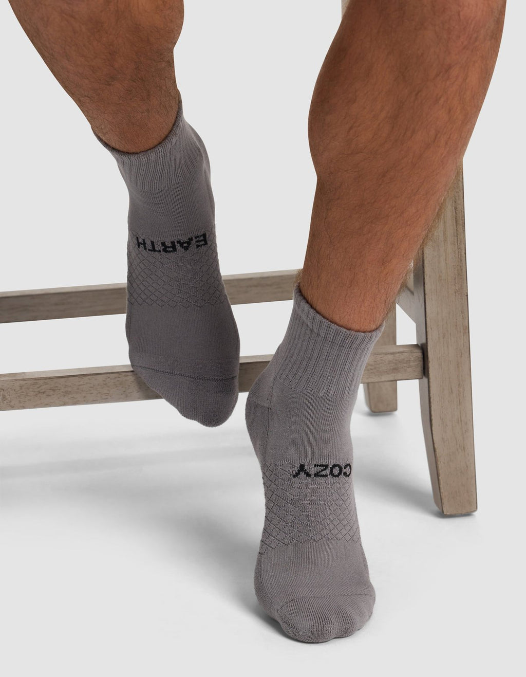 A person sits cross-legged on a wooden stool wearing Cozy Earth Essential Quarter Socks in light gray, featuring "EARTH" and "COZY" in black lettering.|Color:Stone