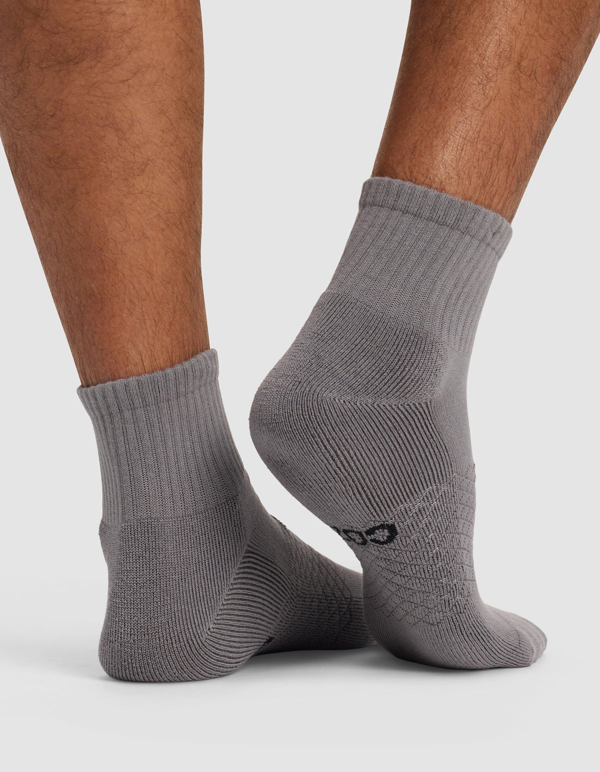 Close-up of a person's lower legs wearing Cozy Earth Essential Quarter Socks in light gray with ribbed cuffs, standing on a white surface. A small black logo appears near the heel of one sock.|Color:Stone