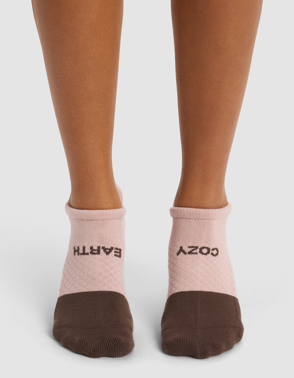A person wears Cozy Earth Essential Ankle Socks in pink and brown; "EARTH" is written on the left sock and "COZY" on the right, both in black letters. The background is plain white.|Color:Cedar