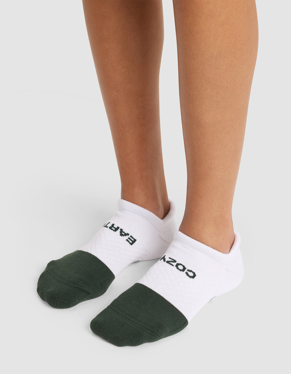 Someone stands on a light surface wearing Cozy Earth Essential Ankle Socks—white with dark green toes and "COZY EARTH" in black lettering on top. |Color:Spruce