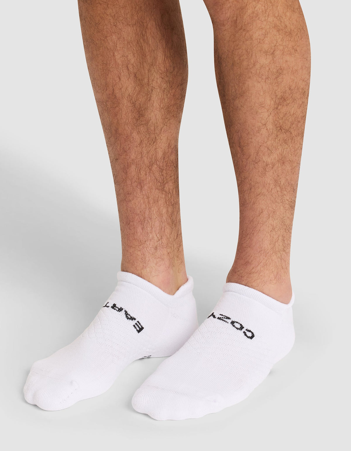 A close-up of a person's lower legs wearing Cozy Earth Essential Ankle Socks in white with black text, standing on a neutral gray background. |Color:White