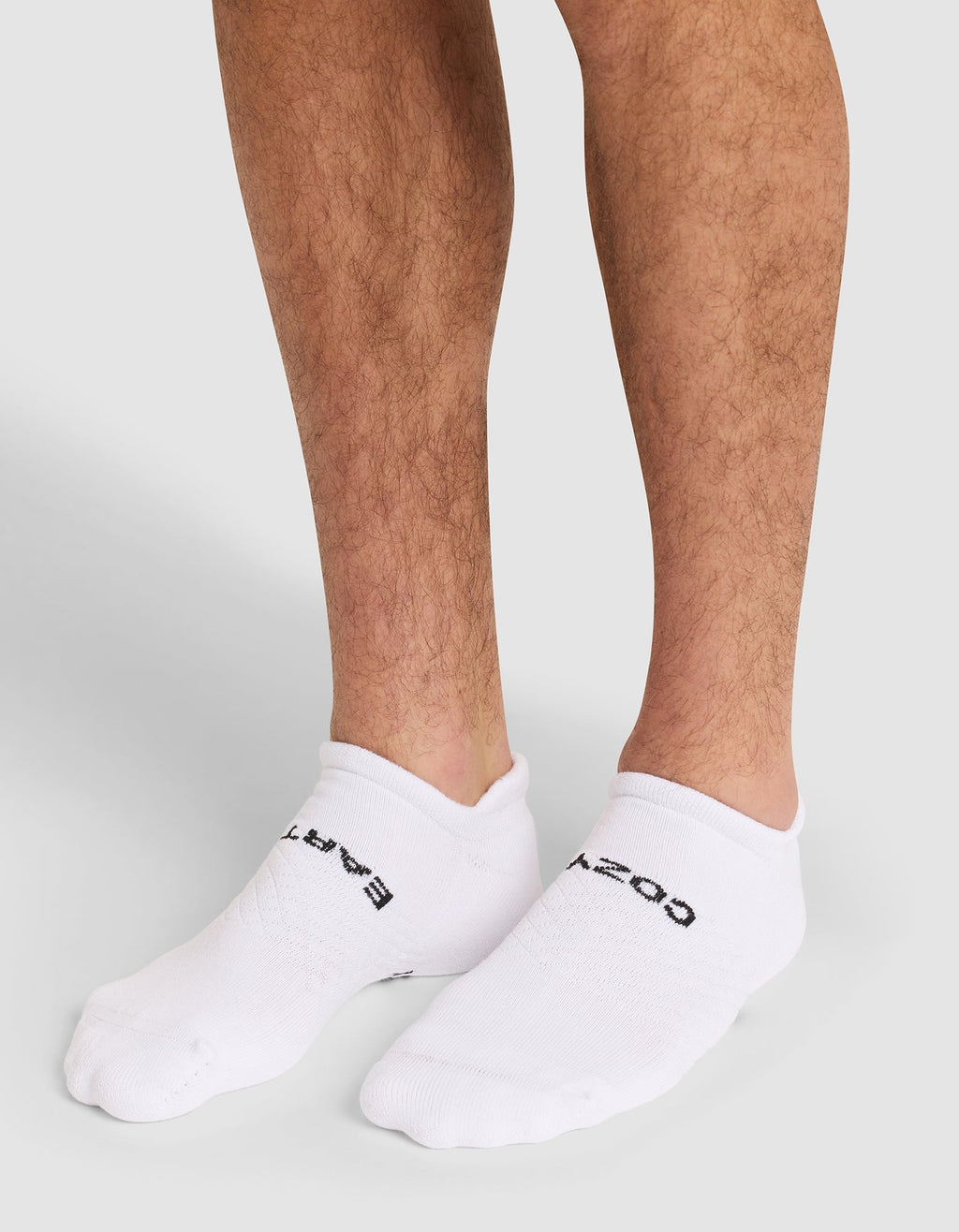 A close-up of a person's lower legs wearing Cozy Earth Essential Ankle Socks in white with black text, standing on a neutral gray background. |Color:White