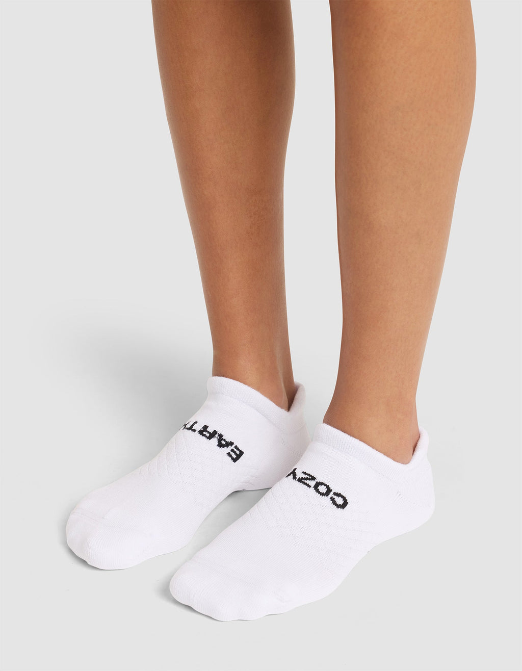 A person wears Cozy Earth Essential Ankle Socks with black text atop, standing on a light gray surface; only their lower legs and feet are shown. |Color:White