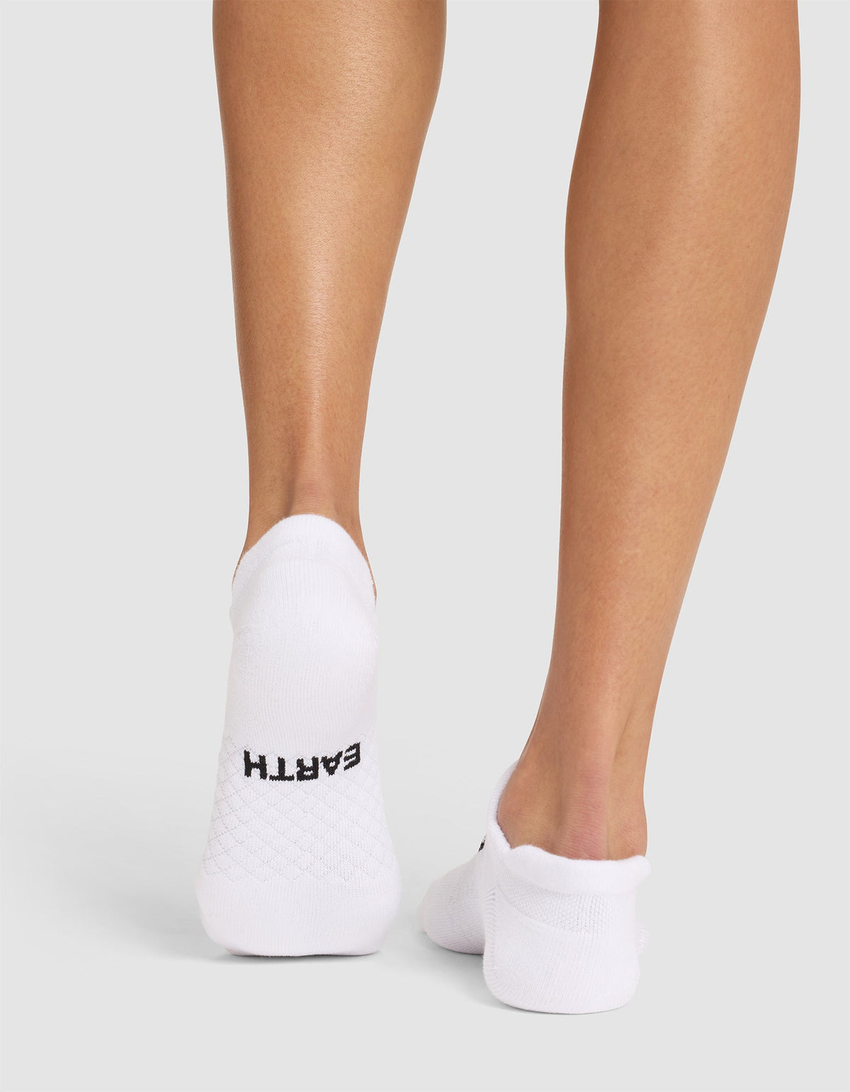 A person stands on a light surface wearing Cozy Earth Essential Ankle Socks. The left sock features "EARTH" in black near the heel. The background is plain, light gray. |Color:White