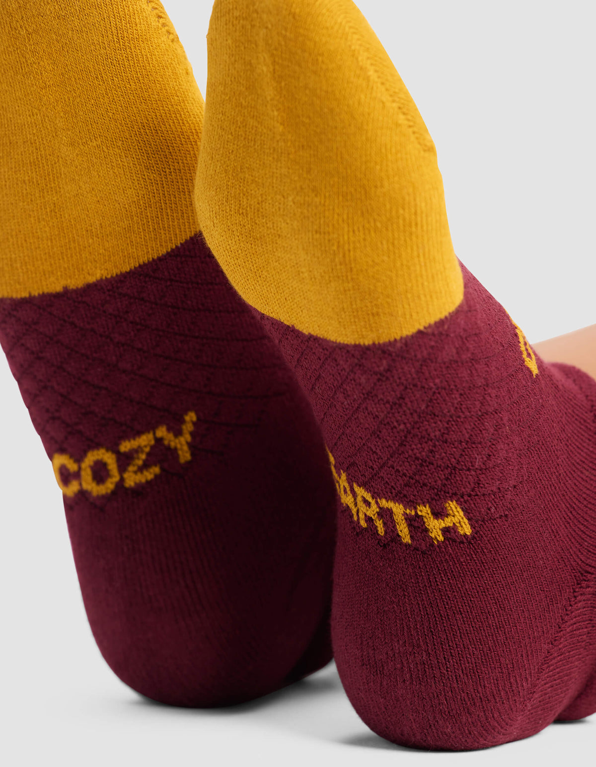 Close-up of two feet wearing Cozy Earth Women's Essential Ankle Socks featuring maroon heels, yellow tops, and "COZY" and "EARTH" in yellow text on each heel. The background is plain light gray.|Color:Burgundy
