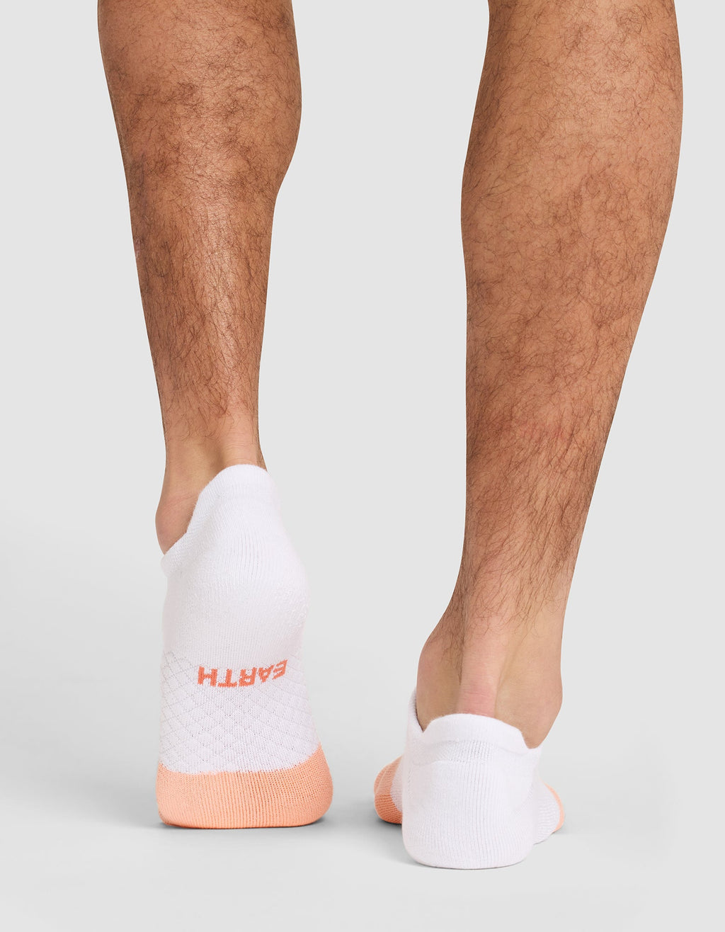 Someone stands on a light gray surface wearing Cozy Earth Essential Ankle Socks—white with light orange accents. "EARTH" appears in orange lettering on the back of the left sock. Only their lower legs and feet are shown. |Color:Cantaloupe