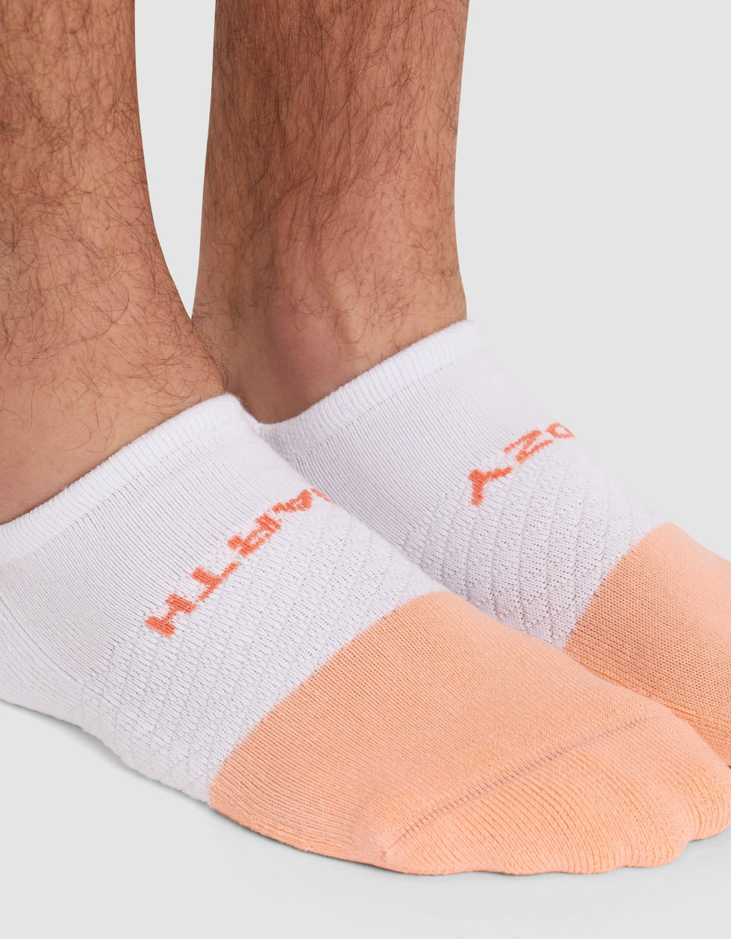 Close-up of a person's lower legs and feet wearing Cozy Earth Essential Ankle Socks in white and light orange, featuring orange text on top. Background is plain light gray. |Color:Cantaloupe