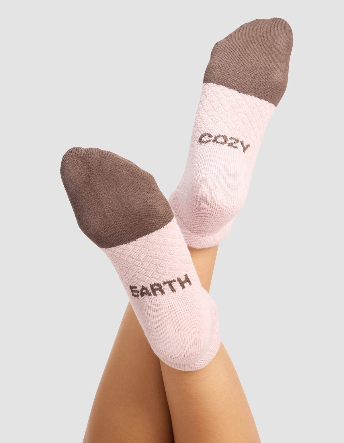 A woman wears Cozy Earth's Women's Essential Ankle Socks in pink with brown toes and heels. "EARTH" is on the left sock, "COZY" on the right. The background is plain light gray.|Color:Cedar