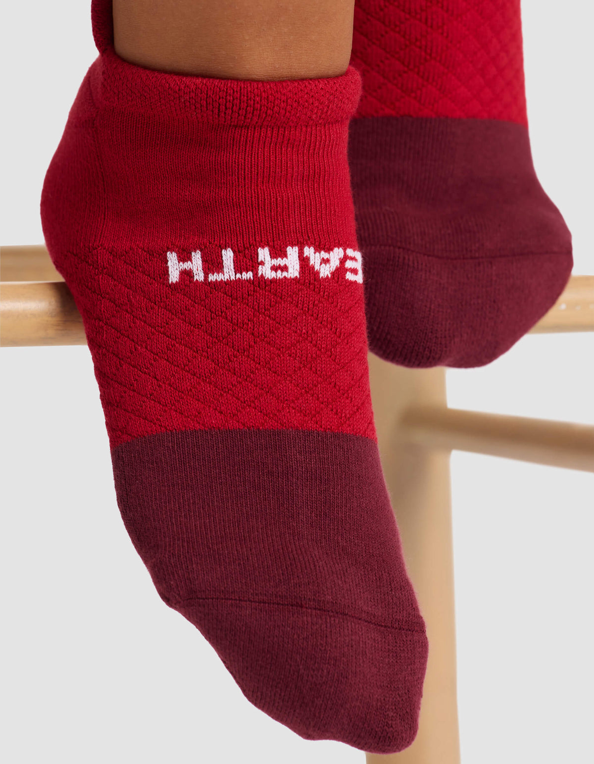 Close-up of feet in Cozy Earth's Women's Essential Ankle Sock—red with darker red toes and heels. "EARTH" in white appears upside down on the sole. The feet rest on a wooden bar.|Color:Crimson