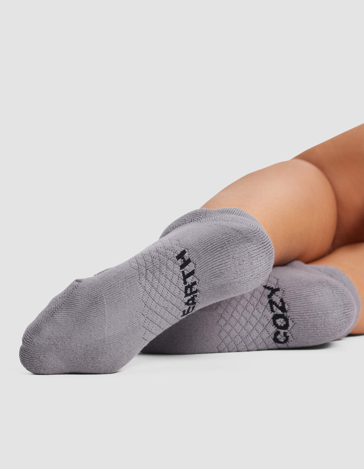 Close-up of a person lying down with feet facing the camera, wearing Cozy Earth Women's Essential Ankle Socks in light gray, featuring "COZY" on one and "CRAFTY" on the other in black letters.|Color:Stone