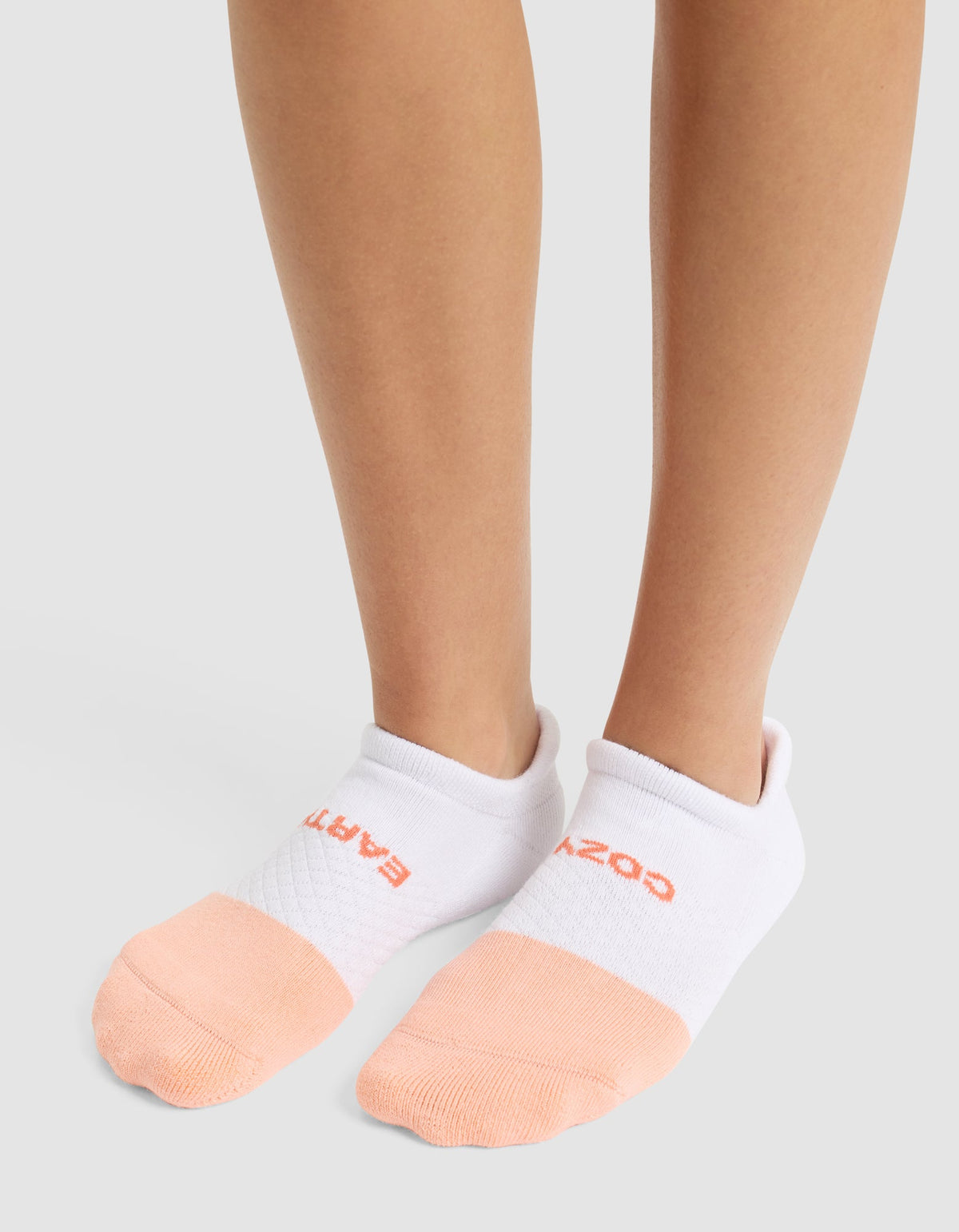 A person wearing Cozy Earth's Essential Ankle Sock—featuring white tops, peach toes, and red text near the cuff—stands on a light gray surface. |Color:Cantaloupe