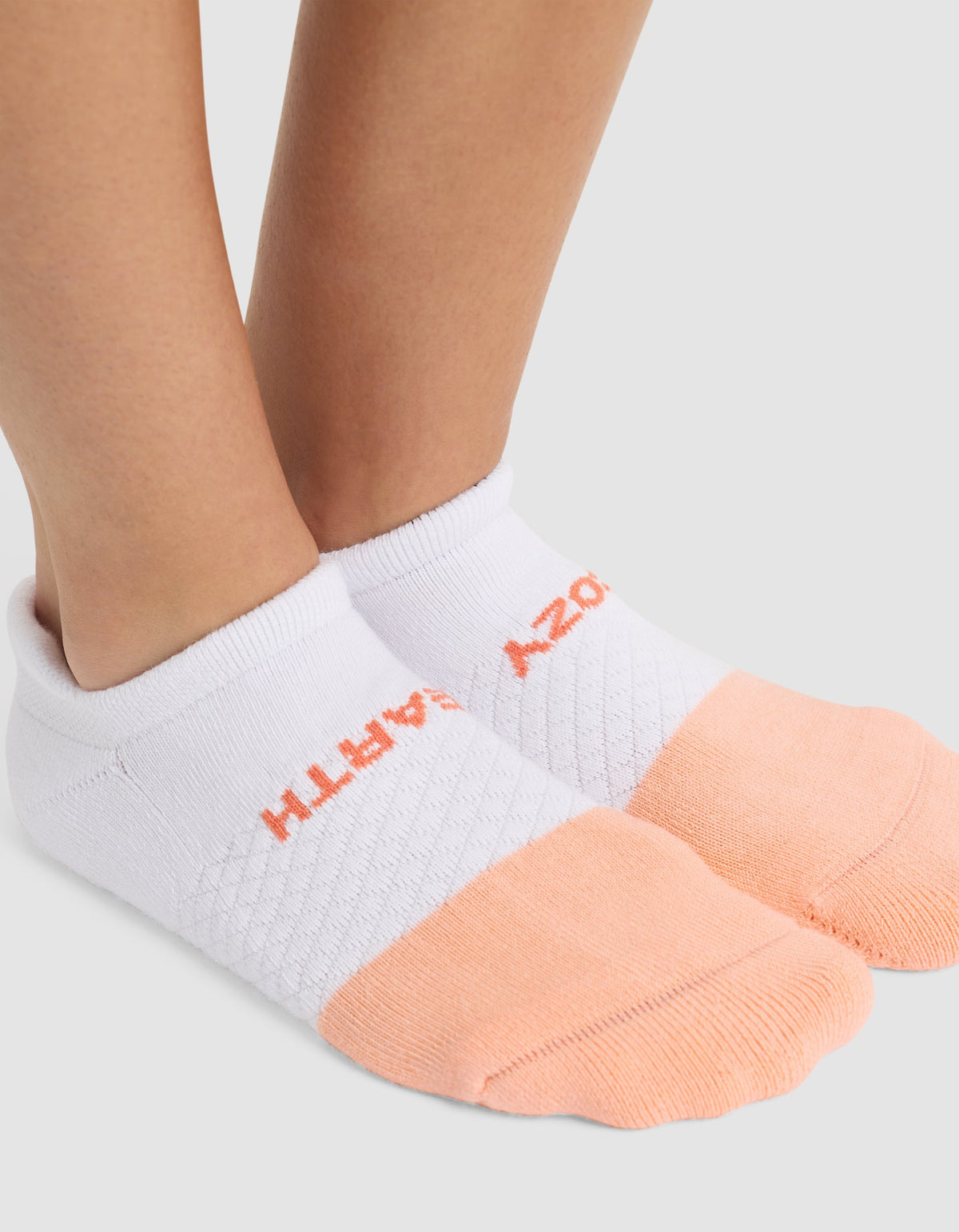 Close-up of a person's feet wearing Cozy Earth's Essential Ankle Sock in white, featuring peach-colored toes and heels. "EARTH" is stitched in orange on the side. |Color:Cantaloupe