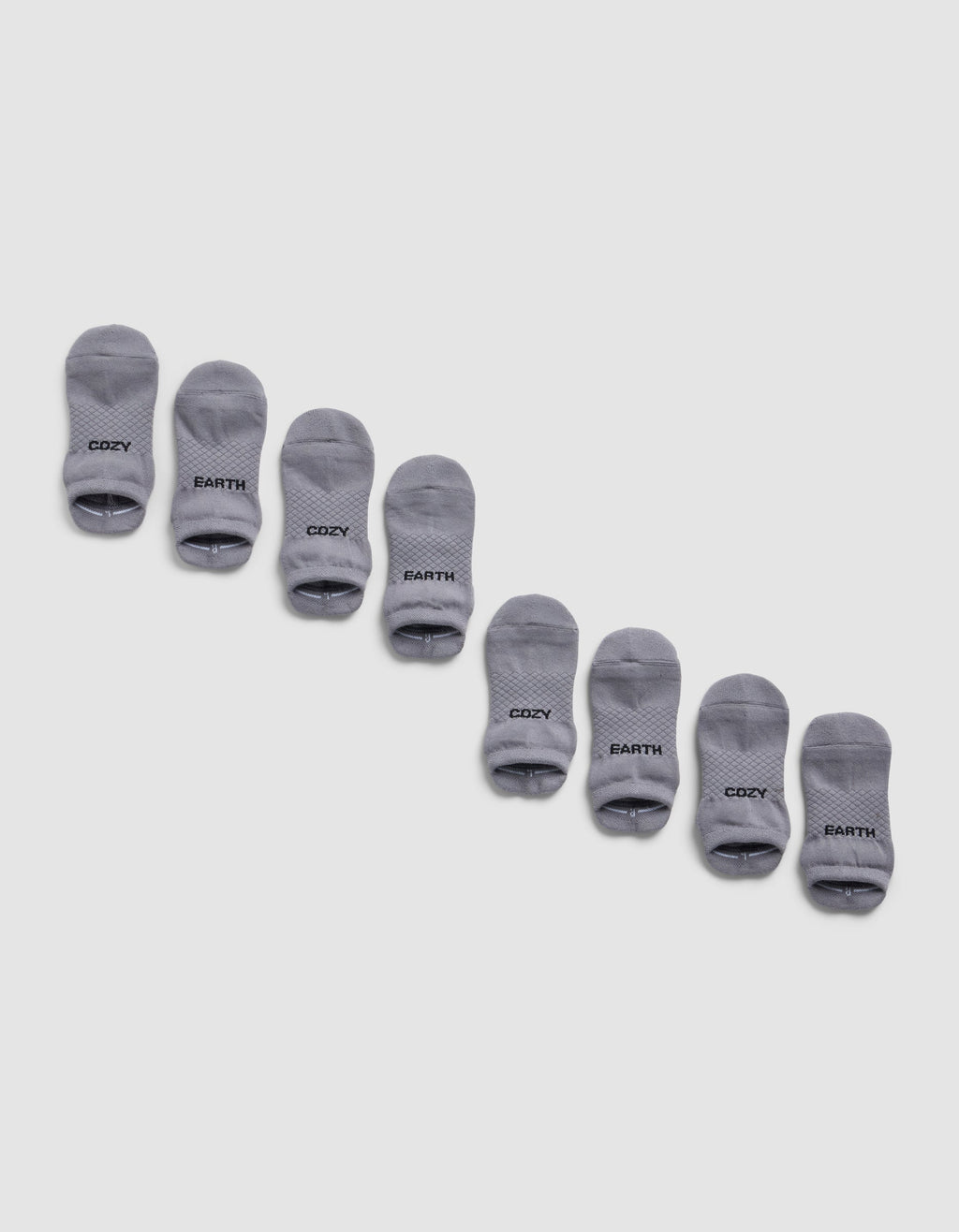 Four pairs of gray Essential Ankle Socks from Cozy Earth are arranged diagonally on a light gray background, each featuring "COZY EARTH" printed in black near the toes.|Color:Stone