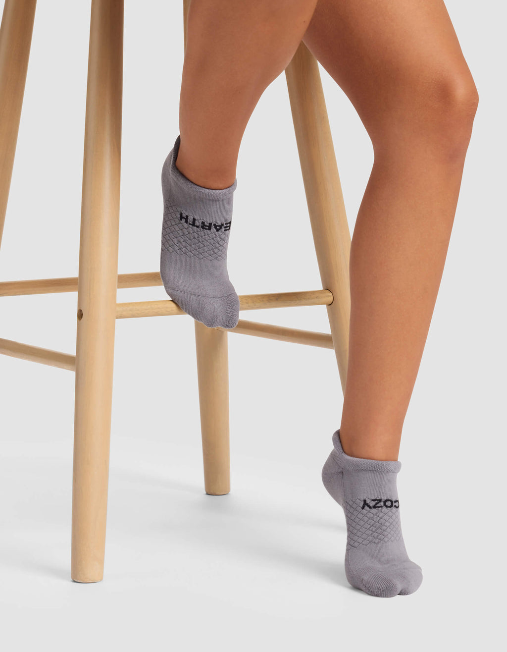 A person wearing Cozy Earth Women's Essential Ankle Socks in gray sits on a light wooden stool, with one foot on a lower rung and the other pointed downward.|Color:Stone