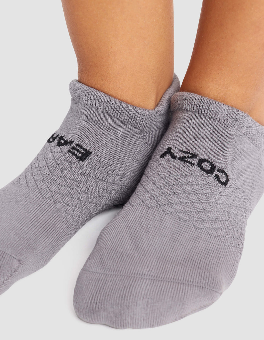 Close-up of feet wearing Cozy Earth Women's Essential Ankle Socks in light gray, featuring black "COZY" on one sock and "BARE" on the other, both clearly visible against a neutral background.|Color:Stone