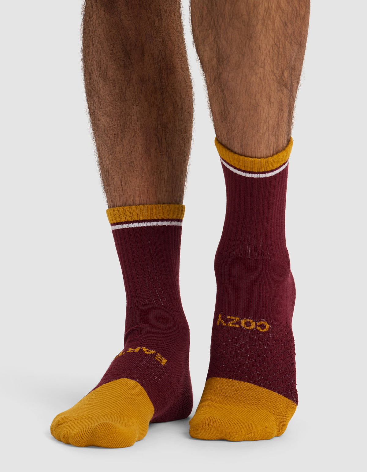 A person wears mismatched Cozy Earth Essential Calf Socks—both maroon with yellow toes and cuffs, but each features different yellow accents and text—shown standing on a white background.|Color:Burgundy