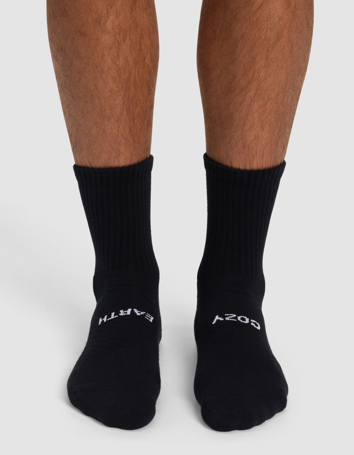 A person wears Cozy Earth Essential Calf Socks in black, featuring "EARTH" on the left and "COZY" on the right sock. Only their lower legs and feet appear against a plain background.|Color:Black