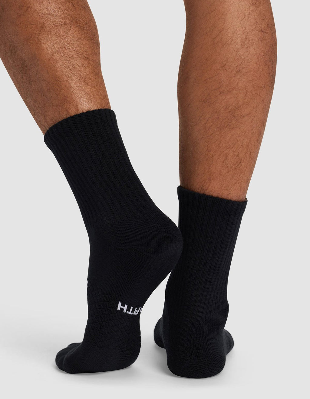 A close-up shows a person wearing Cozy Earth's Essential Calf Sock in black while standing on a light gray surface. "EARTH" is visible near the toes on one sock.|Color:Black