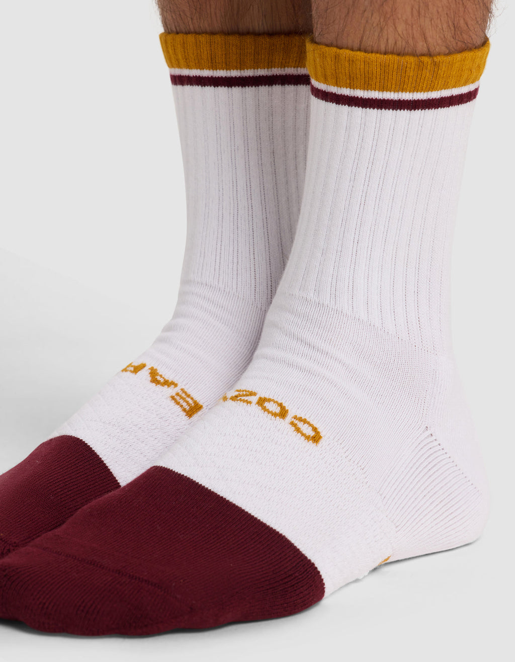 A close-up of a person wearing Cozy Earth's Essential Calf Sock, a white ribbed mid-calf sock with maroon toes and heels, a gold top stripe, and yellow text by the toes, shown as part of the 4-pack set.|Color:Burgundy