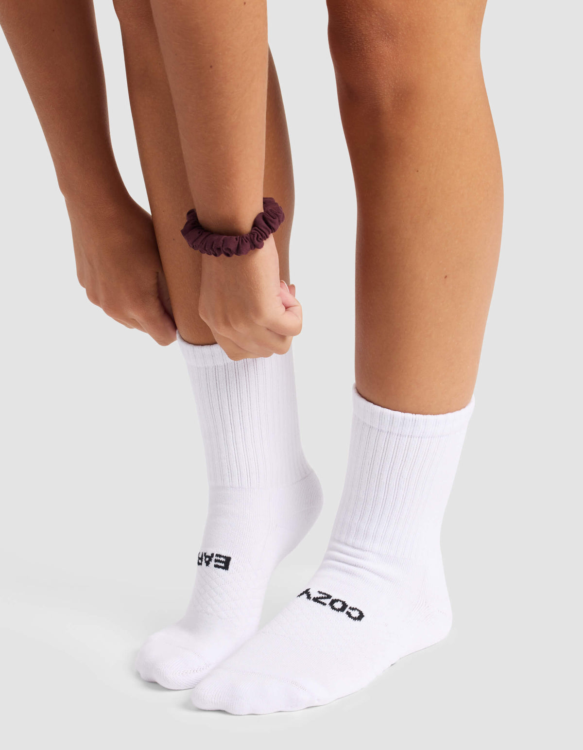 A person wearing Cozy Earth's Women's Essential Calf Sock in white, featuring "COZY" and "EASY" text, adjusts one sock. They have a dark scrunchie on their wrist and stand on a light surface.|Color:White