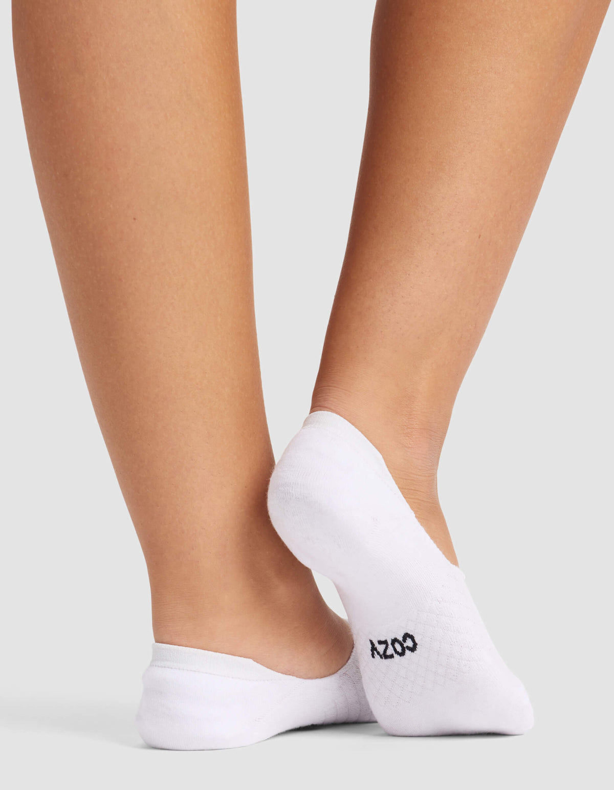 Close-up of a person’s lower legs and feet wearing Cozy Earth Women's Essential No-Show Socks with "COZY" in black on the sole, standing against a plain light gray background.|Color:White