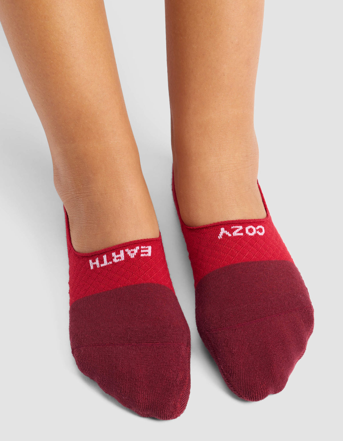 A person stands on a light gray surface wearing Cozy Earth's Women's Essential No-Show Socks in maroon, featuring "EARTH" on the left sock and "COZY" on the right.|Color:Crimson