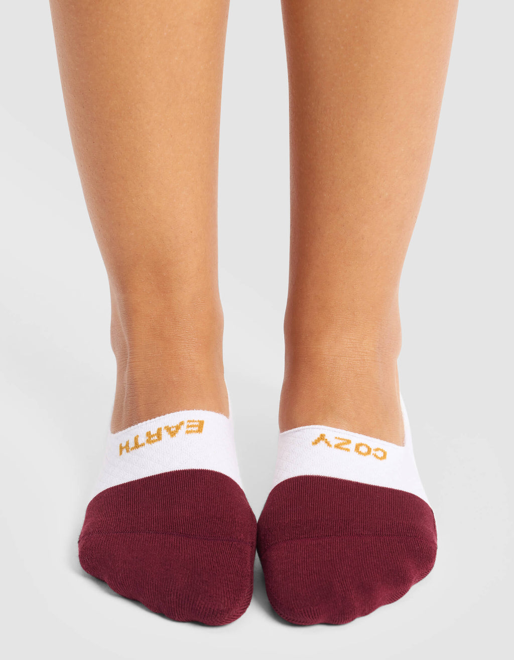 A woman wears Cozy Earth Women's Essential No-Show Socks in white and maroon, featuring "EARTH" on the left and "COZY" on the right sock, both written in yellow across the top.|Color:Burgundy