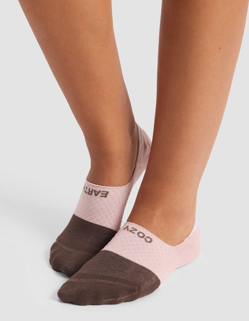 A person stands on a light gray surface wearing Cozy Earth Women's Essential No-Show Socks—low-cut, two-tone with brown toes, pale pink heels, and "COZY" on one sock, "EARTH" on the other.|Color:Cedar