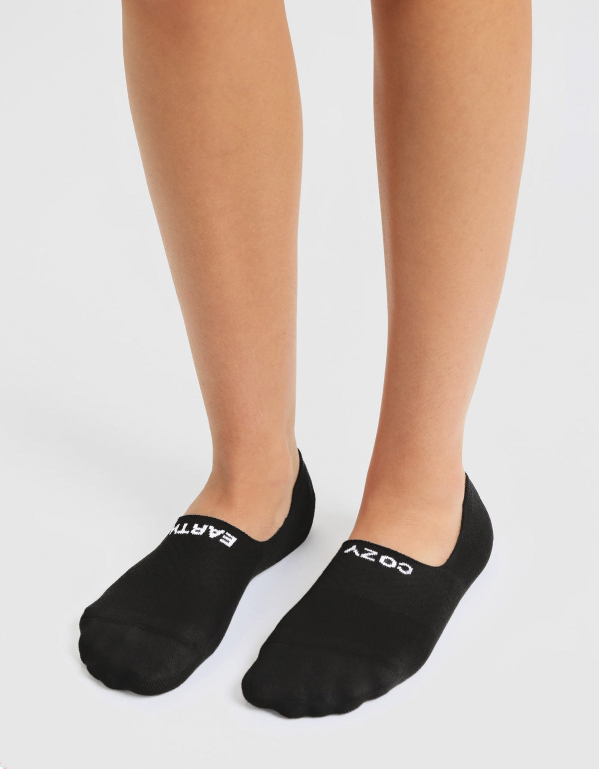 A person stands on a white surface wearing Cozy Earth Women's Essential No-Show Socks in black, with "EARTH" in white on the left sock and "COZY" in white on the right.|Color:Black