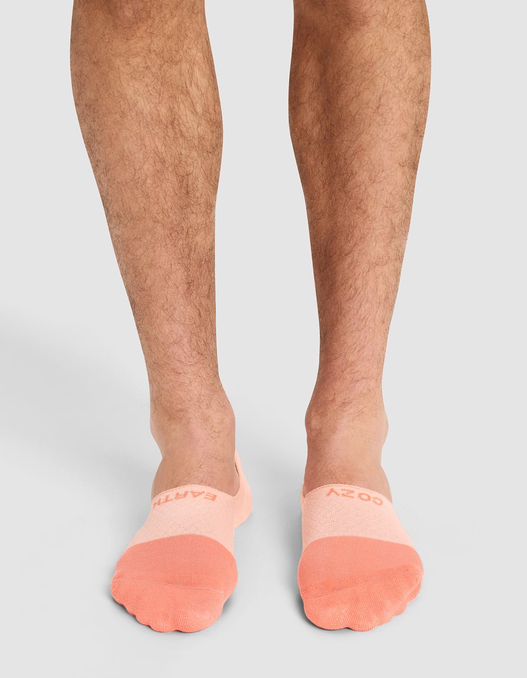 A person wearing Cozy Earth Essential No-Show Socks in light pink with peach toes, featuring text near the toes, stands on a light gray surface. Only their lower legs and feet are visible.|Color:Cantaloupe