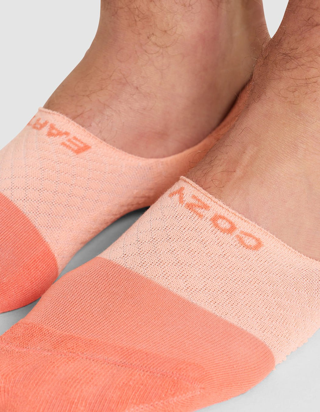 Close-up of two feet wearing Cozy Earth Essential No-Show Socks in light peach and coral with subtle texture. "COZY" is visible on one sock. Background is plain, light gray.|Color:Cantaloupe