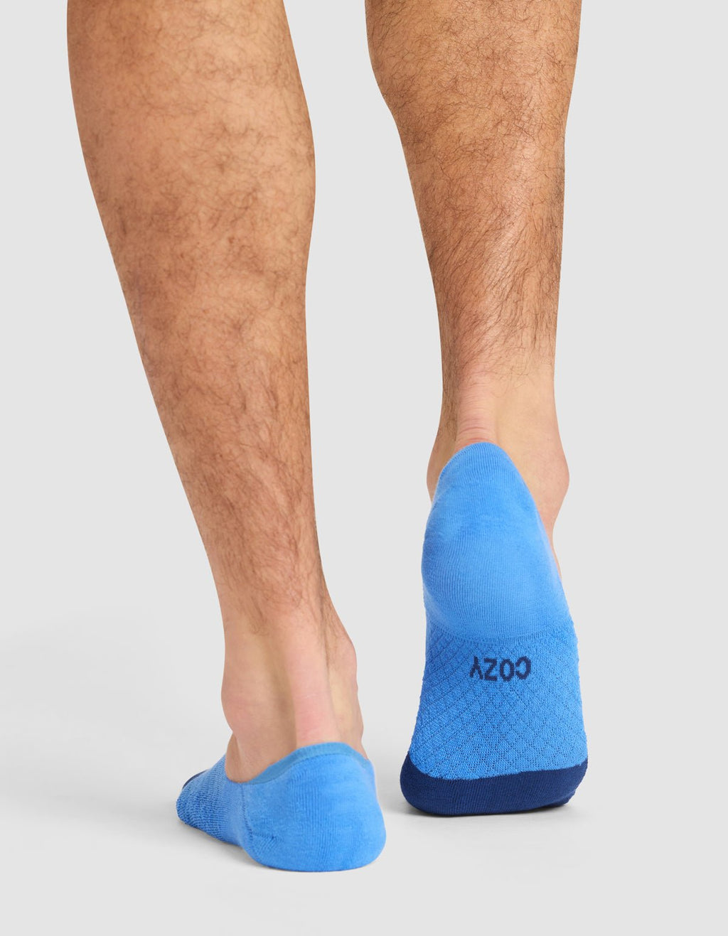 A person stands on a white surface, seen from behind, wearing Cozy Earth Essential No-Show Socks in bright blue. The left sock features "COZY" in black text near the heel.|Color:Harbor