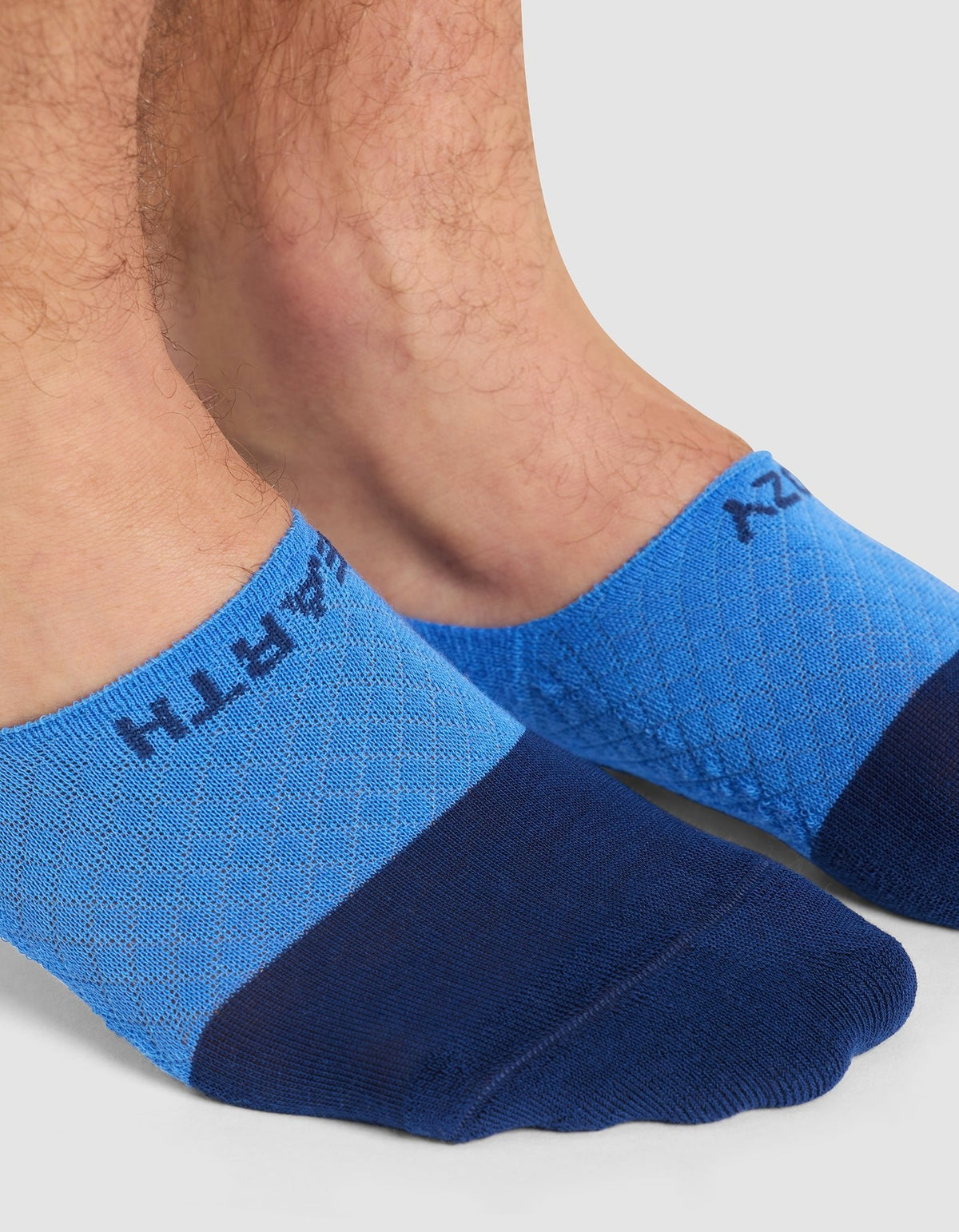 Close-up of two feet wearing Cozy Earth Essential No-Show Socks in blue, featuring a darker blue toe and "EARTH" near the ankle, shown on a light background.|Color:Harbor
