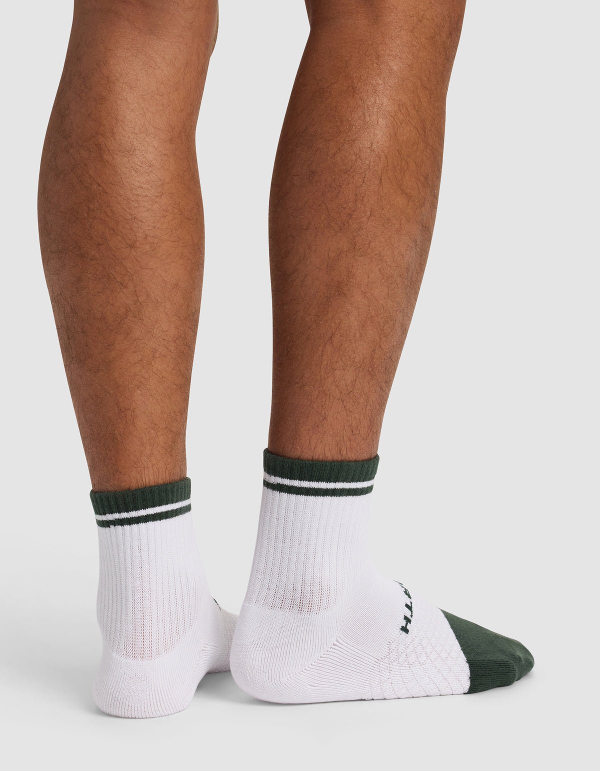 A person stands with their back to the camera, showing only their lower legs and feet in Cozy Earth’s Essential Quarter Sock - Last Chance: white socks with dark green cuffs and toes, ribbed texture, and a subtle sole pattern.|Color:Spruce