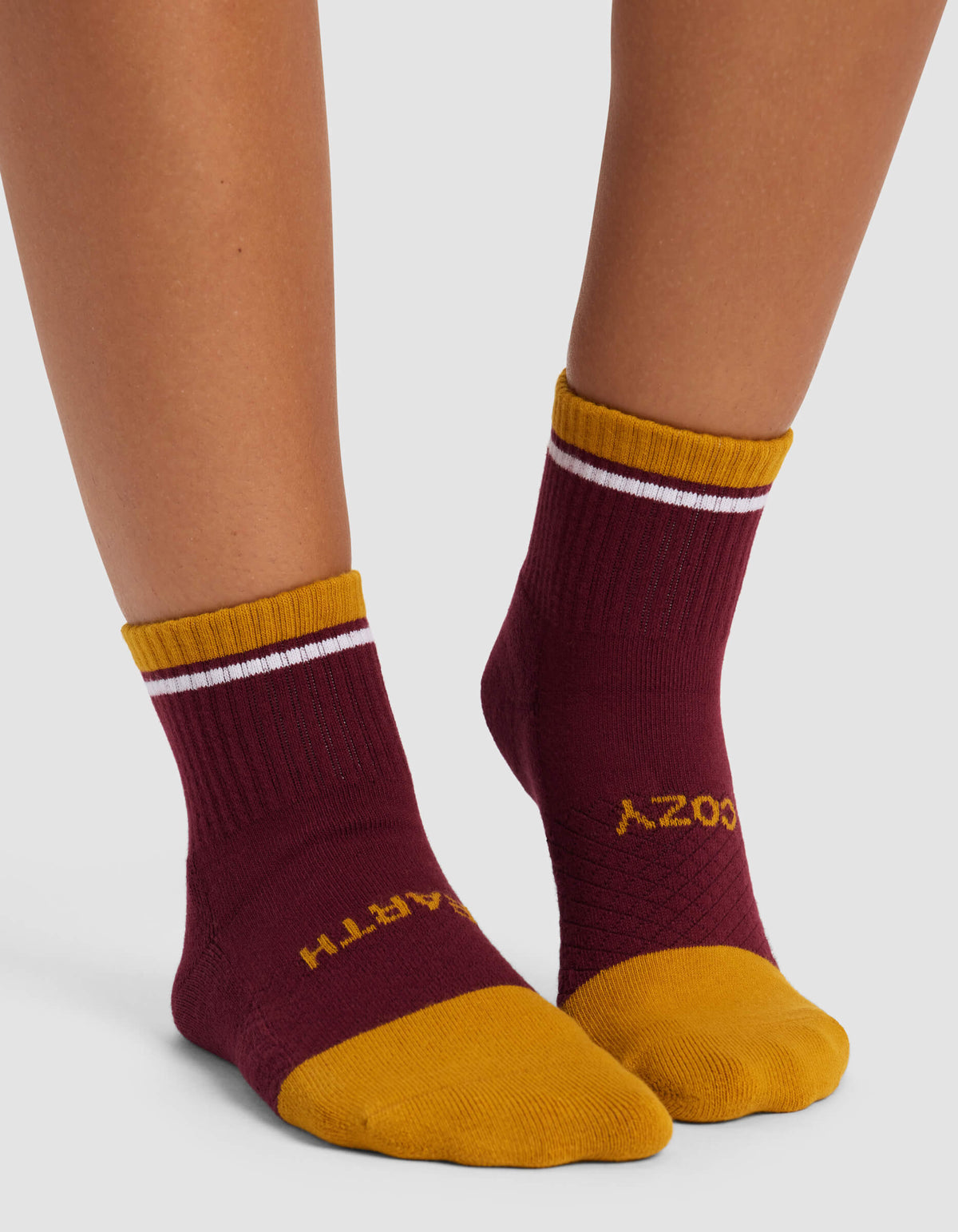 A person wears Cozy Earth Women's Essential Quarter Socks in maroon with gold toes, heels, and cuffs featuring a white stripe; "EARTH" is on the left sock and "COZY" on the right in yellow letters.|Color:Burgundy