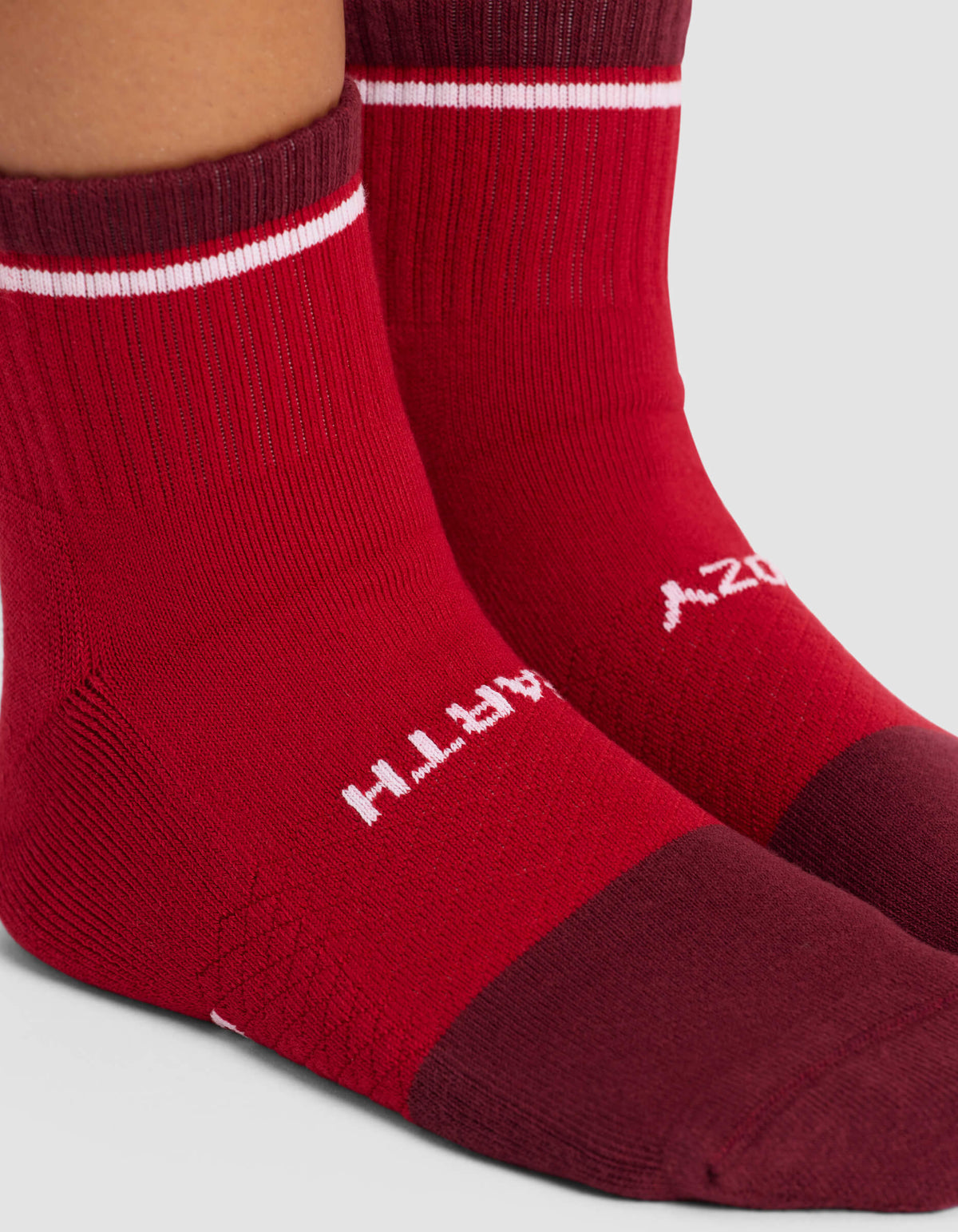 Close-up of two feet wearing Cozy Earth's Women's Essential Quarter Sock in red with a darker maroon toe and heel, a white stripe near the top, and upside-down white "EARTH" text on the side.|Color:Crimson