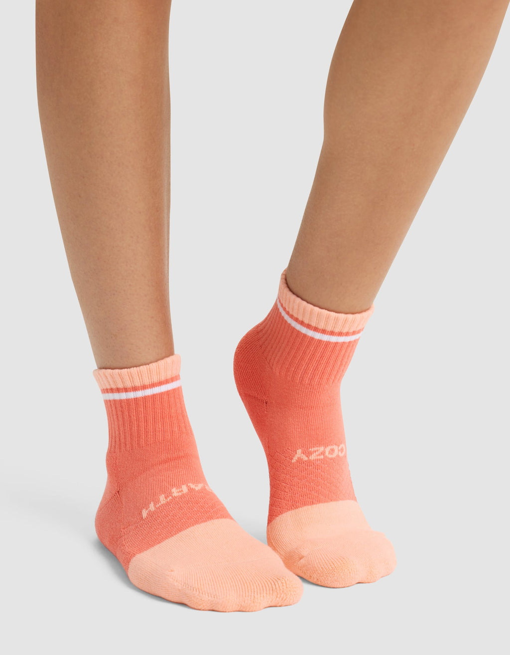 A person wearing Cozy Earth Essential Quarter Socks in peach and coral with white stripes and white front lettering, standing on a light gray surface. |Color:Cantaloupe