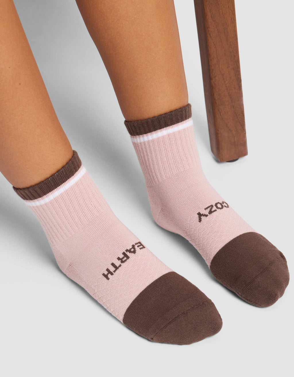 A person wearing Cozy Earth Women's Essential Quarter Socks in pink with brown toes and cuffs, featuring “COZY” on the right foot and “EARTH” on the left, sits near a wooden chair.|Color:Cedar