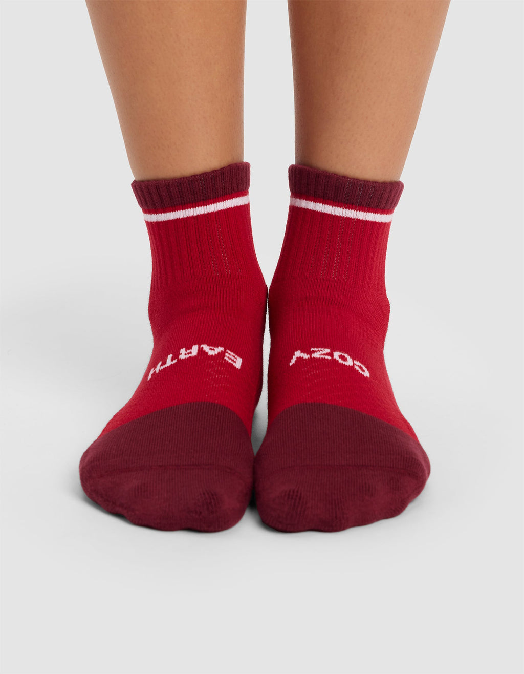 A pair of feet wearing Cozy Earth Essential Quarter Socks in red, featuring darker red toes and cuffs, white stripes near the top, and partially visible white text. The feet stand on a light grey surface.|Color:Crimson