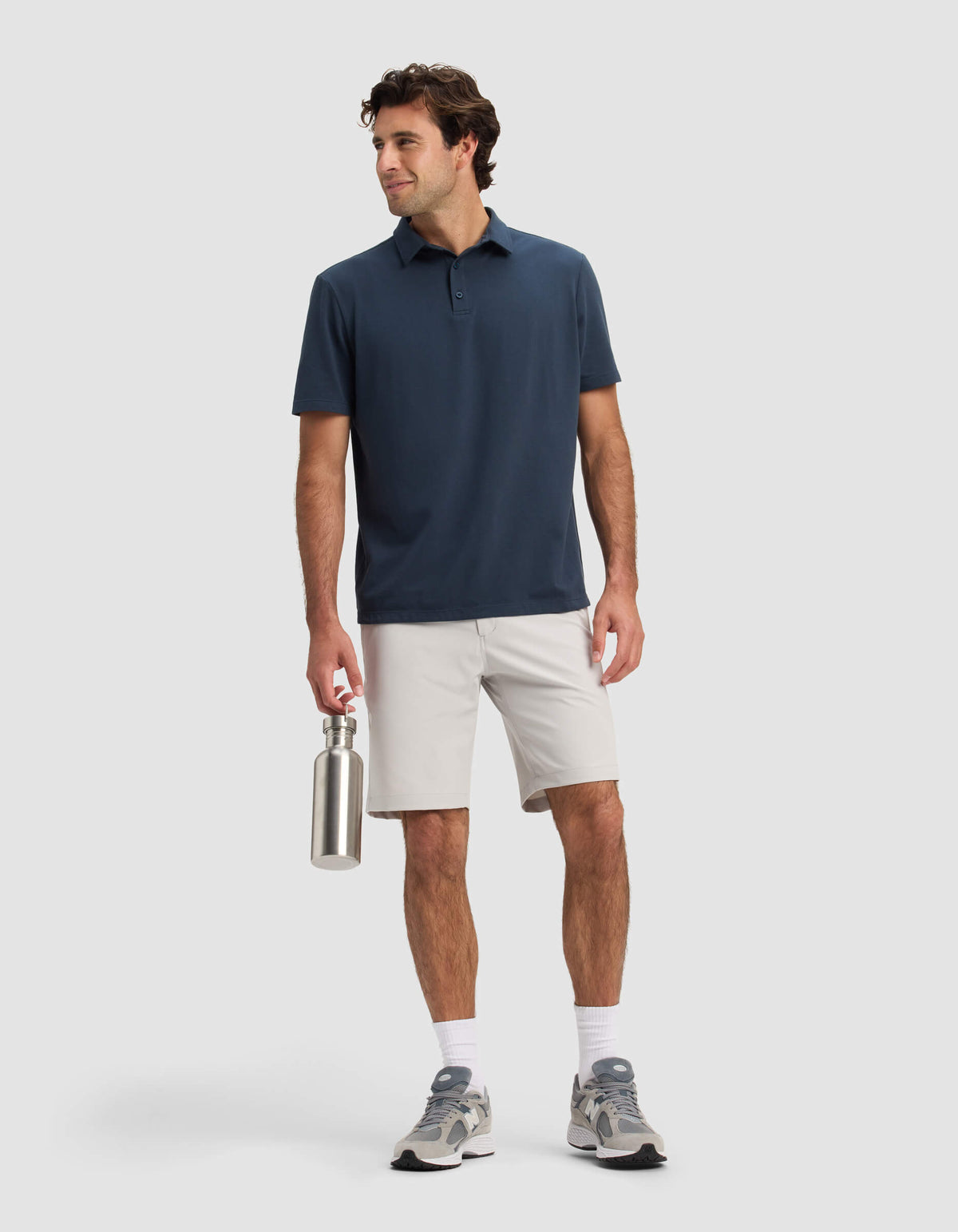 A man stands smiling against a plain background, wearing the Cozy Earth Men's Everywhere Short in light gray, paired with a navy polo, white socks, gray sneakers, and holding a metal water bottle.|Color:Dew