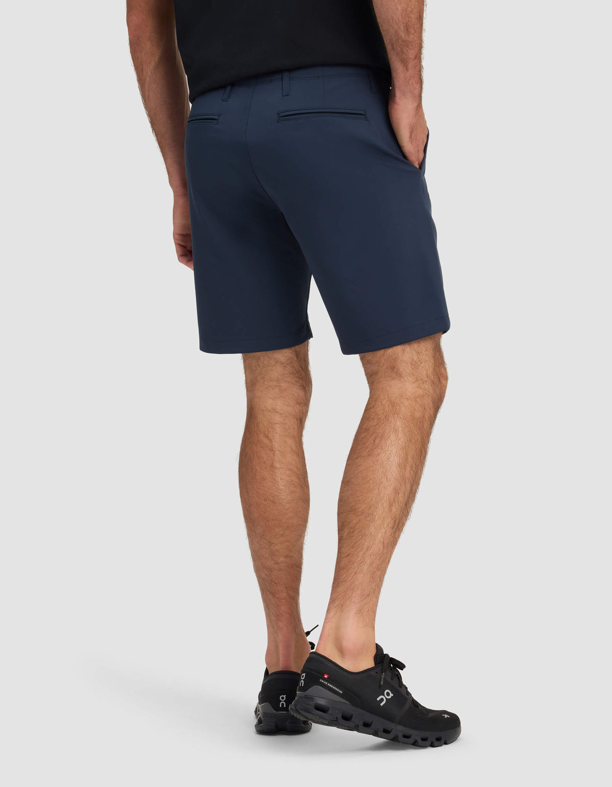 A person wearing Cozy Earth's Men's Everywhere Short in dark blue, with a black shirt and black athletic shoes, stands facing away from the camera with their left hand in their pocket against a plain light background.|Color:Eclipse