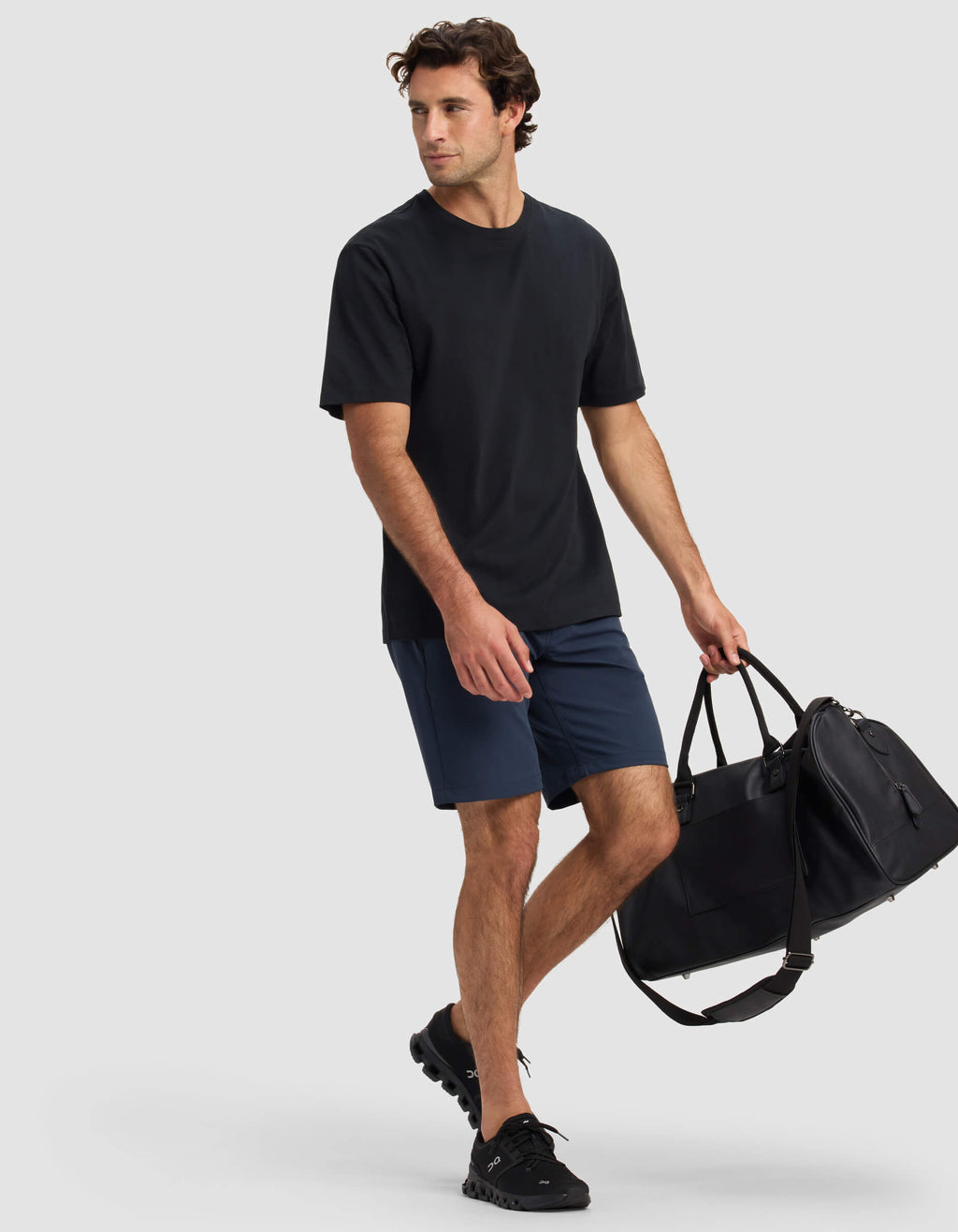 A man wearing Cozy Earth's Men's Everywhere Short in navy and a black t-shirt walks with a large black duffel bag with a shoulder strap and black sneakers against a plain light background.|Color:Eclipse