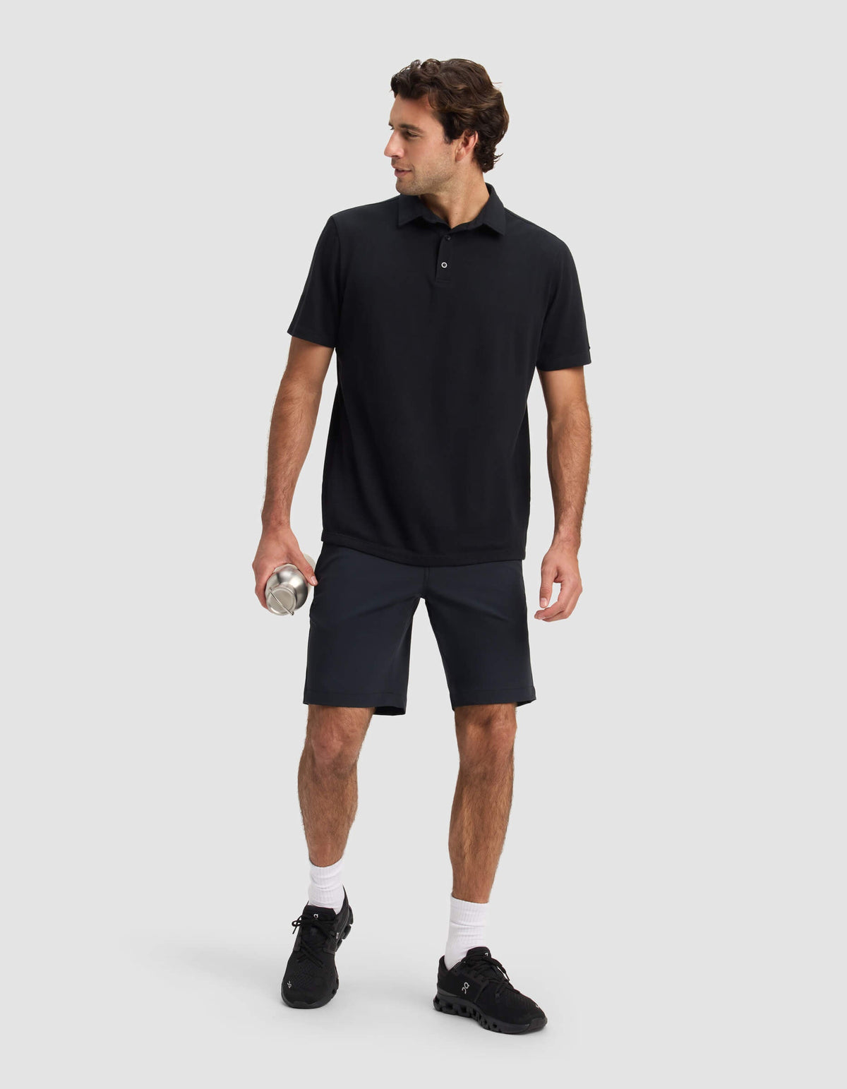 A man stands against a light gray background, wearing Cozy Earth's Men's Everywhere Short in black, paired with a black polo shirt, black sneakers, and white socks. He holds a silver water bottle and looks to his left.|Color:Jet Black