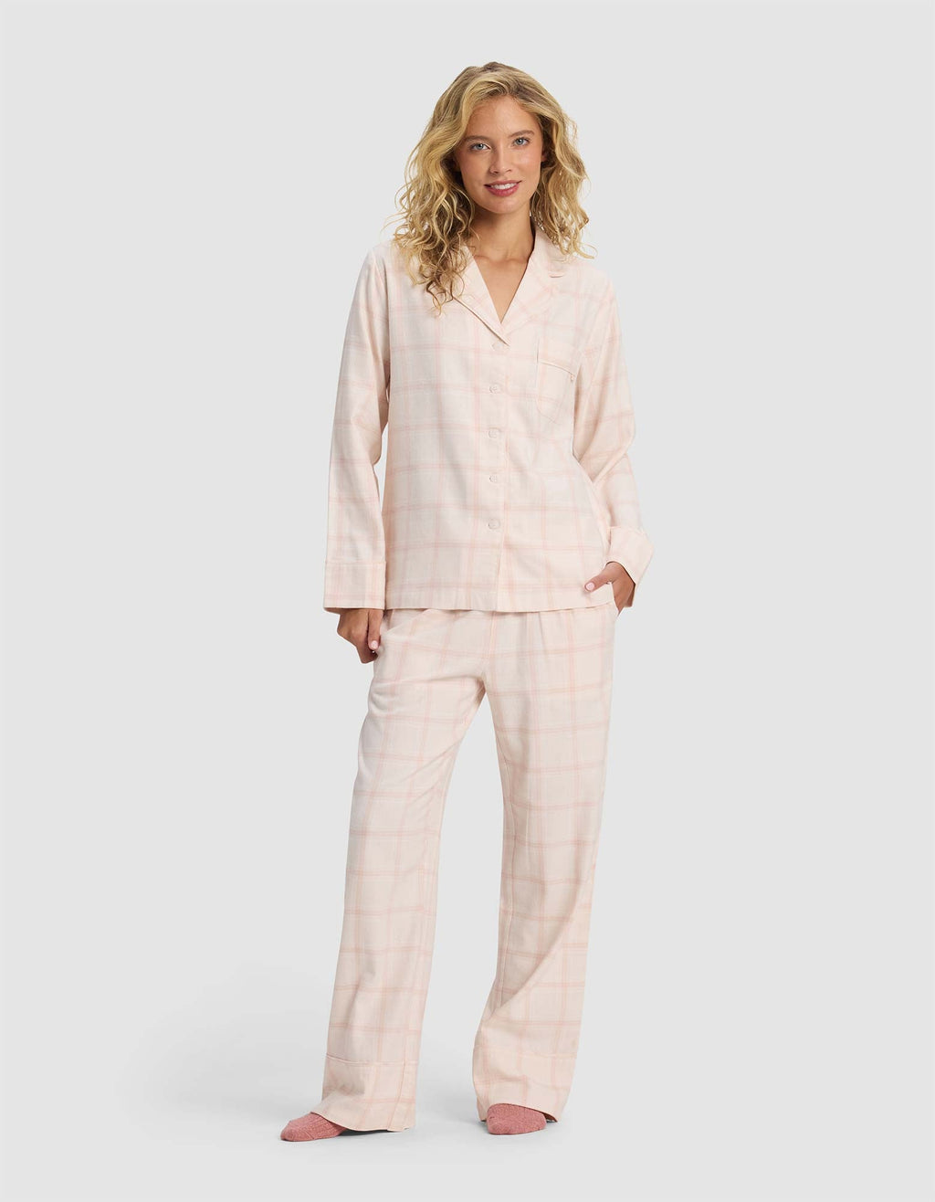 A woman with long blonde hair wears the Cozy Earth Women's Farmhouse Long Sleeve Pajama Top, styled in light pink plaid, paired with pink slippers. She stands against a plain light gray background, smiling slightly with one hand in her pocket.|Color:Quartz Plaid