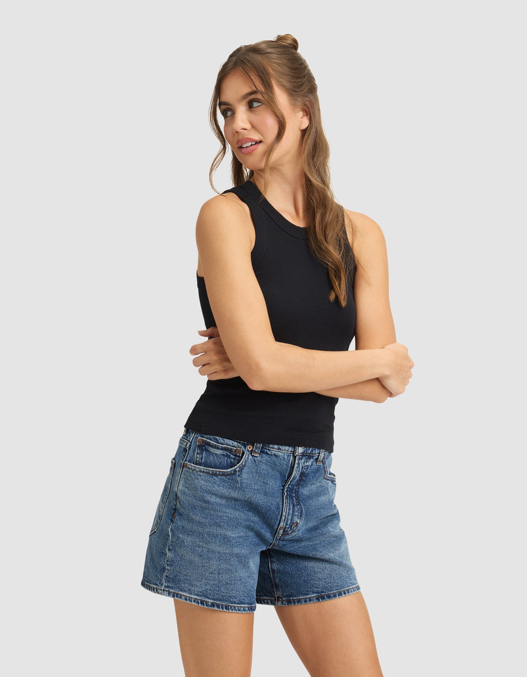 A woman with long brown hair models the Cozy Earth Women's Fine Ribbed Tank in black, paired with blue denim shorts. She stands with arms crossed, looking left against a plain light gray background.|Color:Jet Black