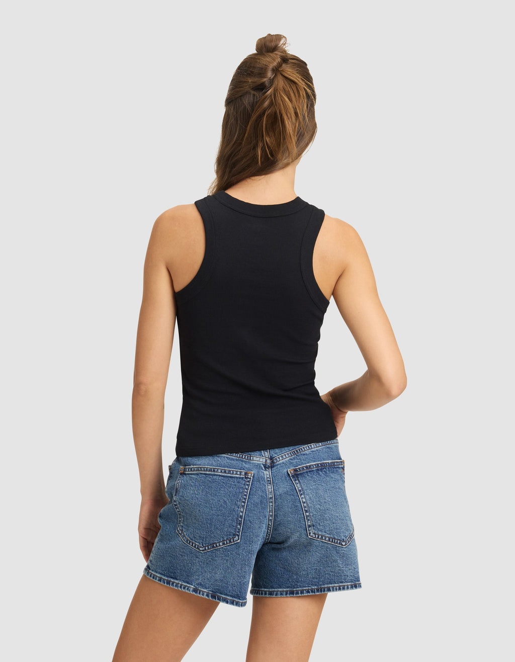 A woman with light brown hair wears the Cozy Earth Women's Fine Ribbed Tank in black, paired with blue denim shorts. She stands facing away from the camera, hand on hip, against a plain light gray background.|Color:Jet Black