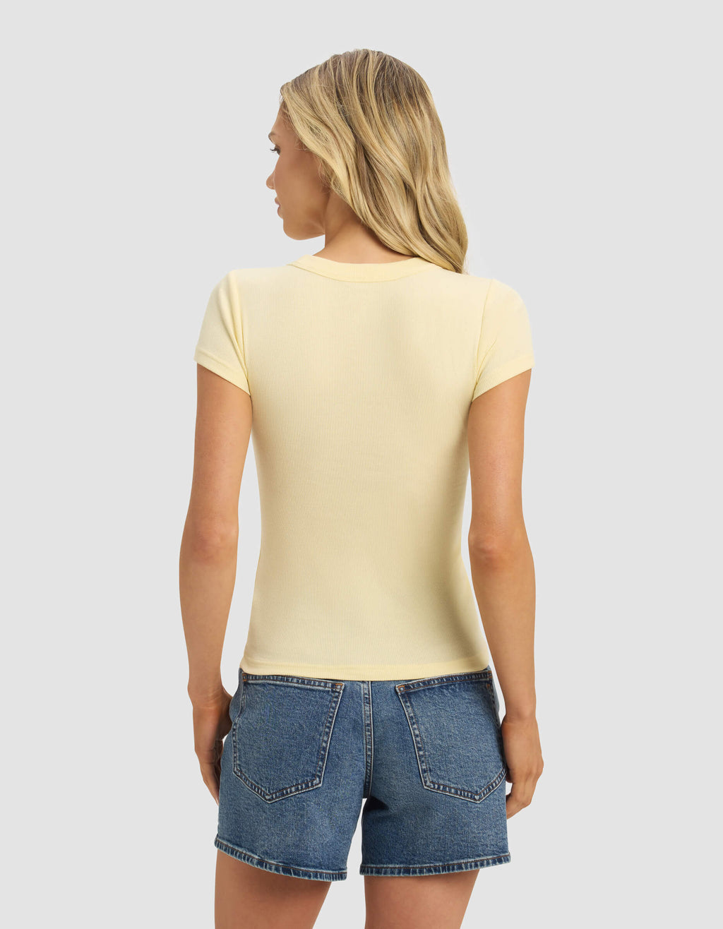 A woman with long blonde hair is seen from behind wearing the Cozy Earth Women's Fine Ribbed Tee in light yellow and blue denim shorts, set against a plain light gray background.|Color:Meringue