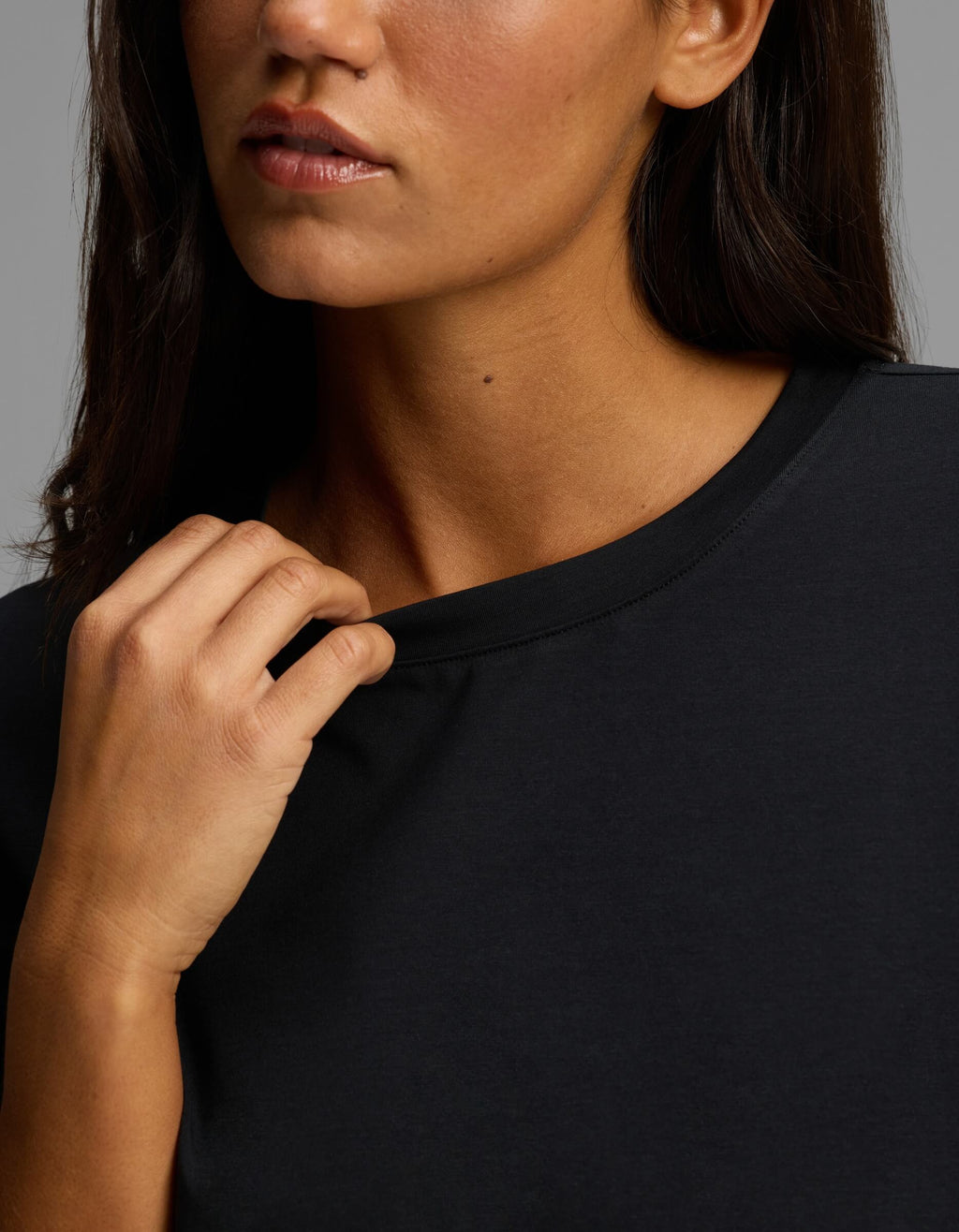 A woman with medium skin tone and straight dark brown hair wears the Cozy Earth Women’s First Layer Boyfriend Sleep Tee in black, gently pulling at its neckline with her right hand. The background is neutral gray, and only the lower half of her face is visible.|Color:Jet Black