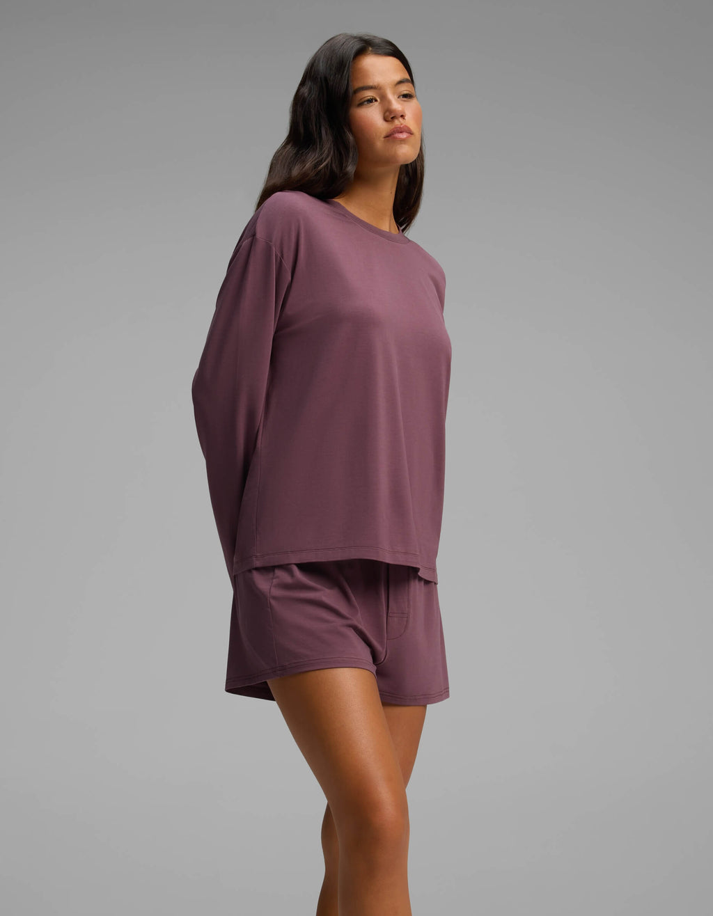 Dressed in the Cozy Earth Women’s First Layer Long Sleeve Sleep Tee & Boxer Short Set, a woman stands against a plain gray background with her hands behind her back, gazing slightly upward.|Color:Fig