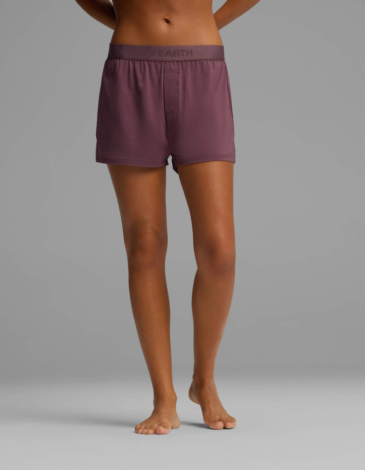 A person stands barefoot on a gray background, wearing mauve boxer shorts from the Cozy Earth Women’s First Layer Pajama Short Sleeve Tee & Boxer Short Set, featuring a waistband with the brand name “COZY EARTH.” Only the lower torso and legs are visible.|Color:Fig